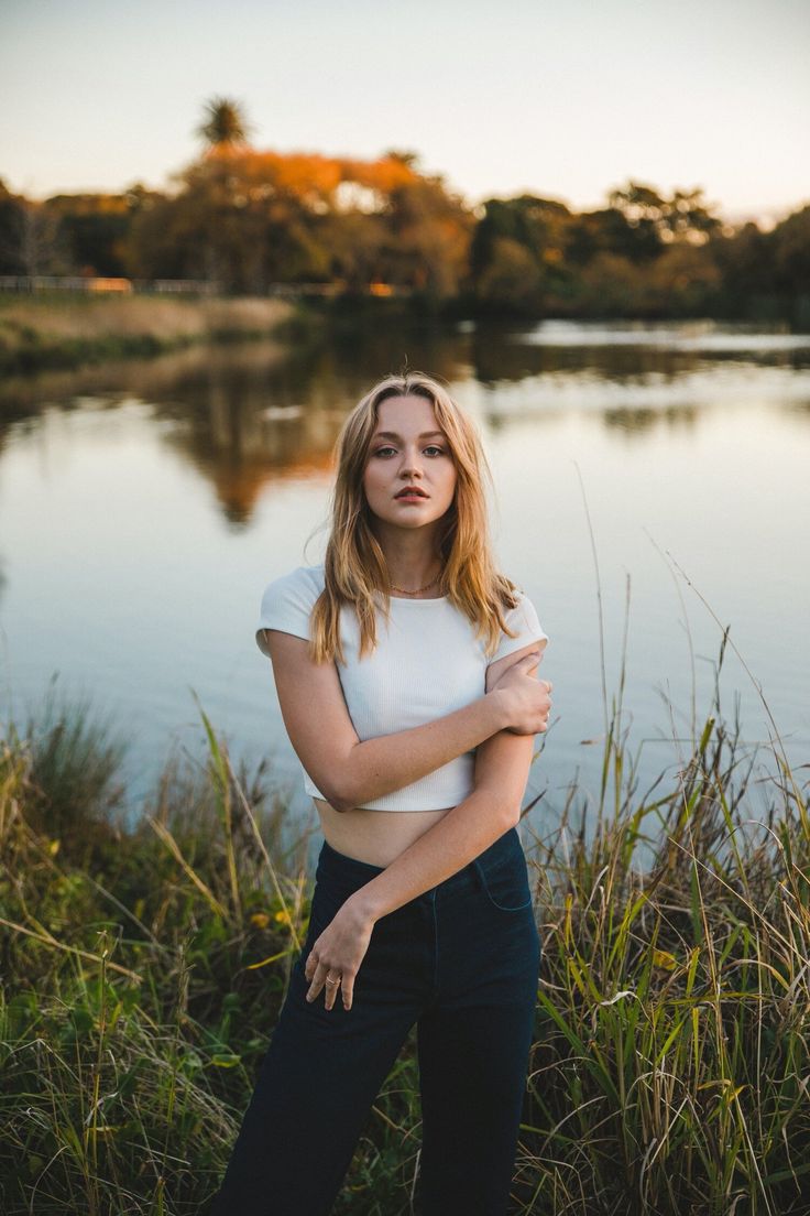 a woman standing in front of a body of water with her arms crossed and looking at the camera