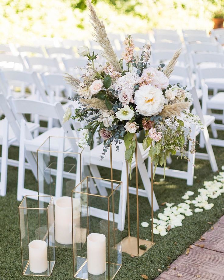 an outdoor ceremony setup with white chairs and tall vases filled with flowers, candles and greenery