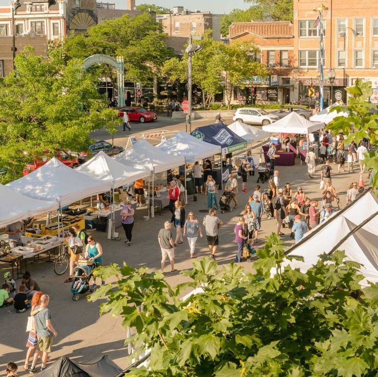 people walking around an outdoor market with tents set up in the middle and buildings in the background