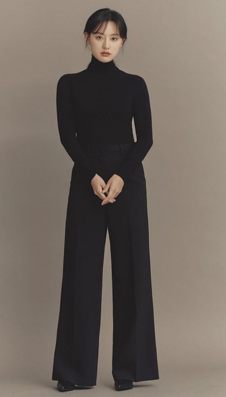 a woman standing in front of a gray background wearing black pants and a turtle neck sweater