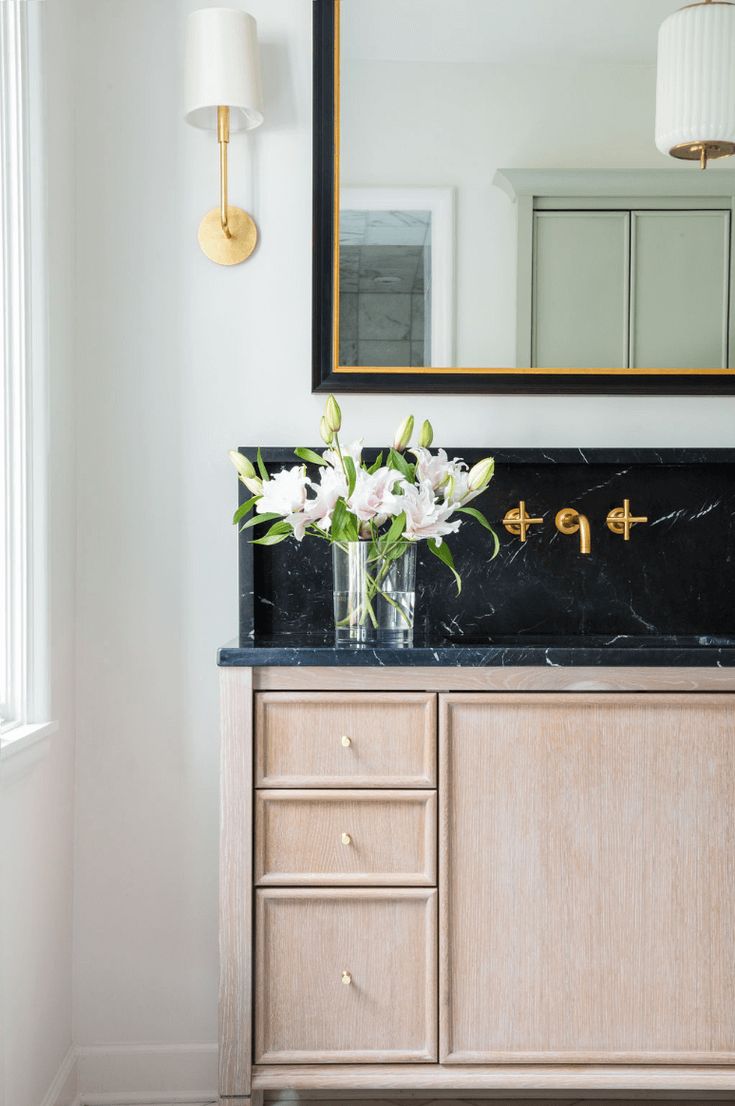 a bathroom vanity with marble counter top and gold faucet, white flowers in glass vase