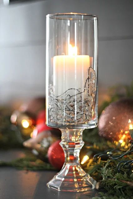 a lit candle in a clear glass on a table with christmas decorations and baubles