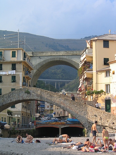 some people are laying on the sand and one is holding a surfboard in front of an old bridge