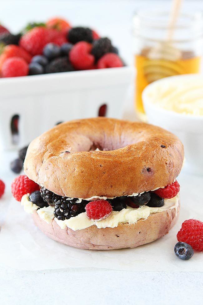 a bagel sandwich with berries, cream cheese and blueberries next to some fruit