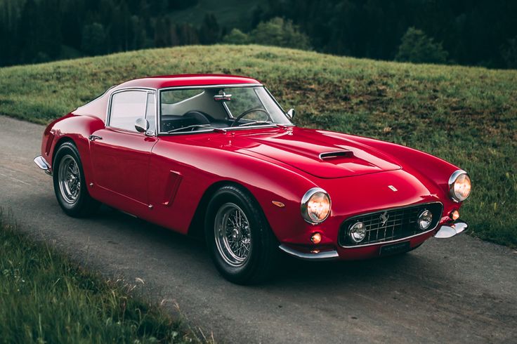 an old red sports car driving down a road in front of some grass and trees