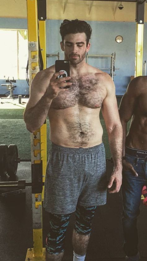 a shirtless man taking a selfie in the gym with his cell phone while standing next to another man