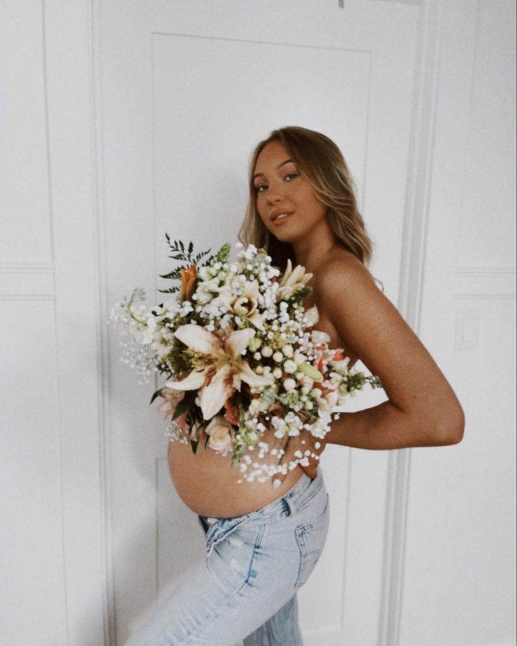 a pregnant woman holding a bouquet of flowers in her stomach while leaning against a wall