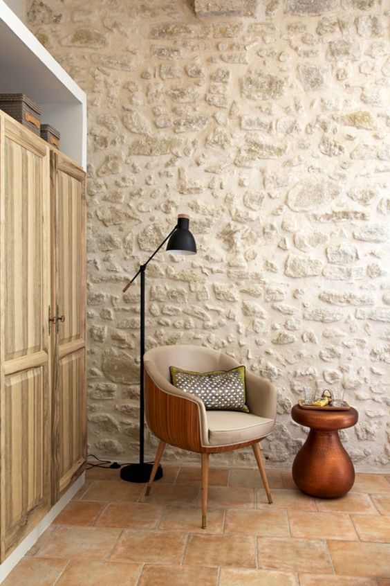 a living room with a chair, lamp and cupboards in front of a stone wall