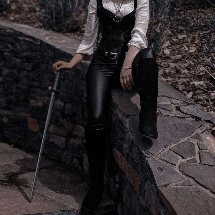 a woman dressed in black and white holding a cane sitting on a stone wall with trees behind her