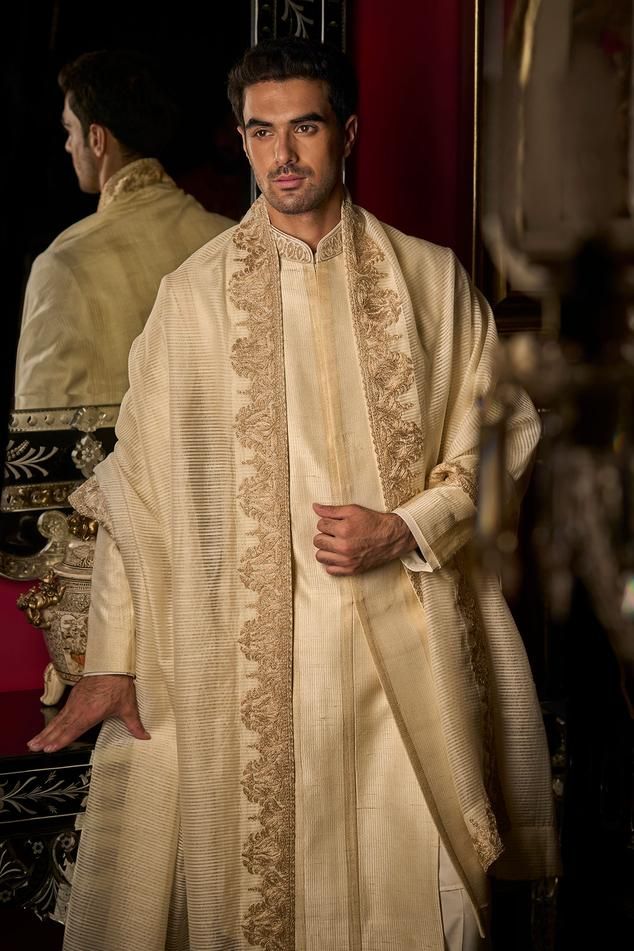 Cream raw silk sherwani with all over striped running stitch line thread work, floral gold zari and dori embroidered mandarin collar. Paired with cotton silk kurta and straight fit trouser.
Components: 3
Pattern: Embroidered
Type Of Work: Thread and Zari Dori Work
Neckline: Mandarin Collar
Sleeve Type: Full Sleeves
Fabric: Sherwani: Raw Silk, Cotton Silk
Color: Cream
Other Details: 
Patch pocket
Side slits
Sherwani Closure: Front concealed placket
Note:
The footwear and dupatta worn by the model Luxury Raw Silk Men's Bandhgala, Luxury Men's Salwar Kameez With Mandarin Collar, Luxury Men's Bandhgala With Mandarin Collar, Luxury Men's Sherwani With Mandarin Collar, Luxury Designer Men's Sherwani, Luxury Men's Sequined Sherwani, Luxury Red Men's Sherwani, Luxury Designer Bandhgala With Mandarin Collar, Luxury Dori Work Sets For Ceremony