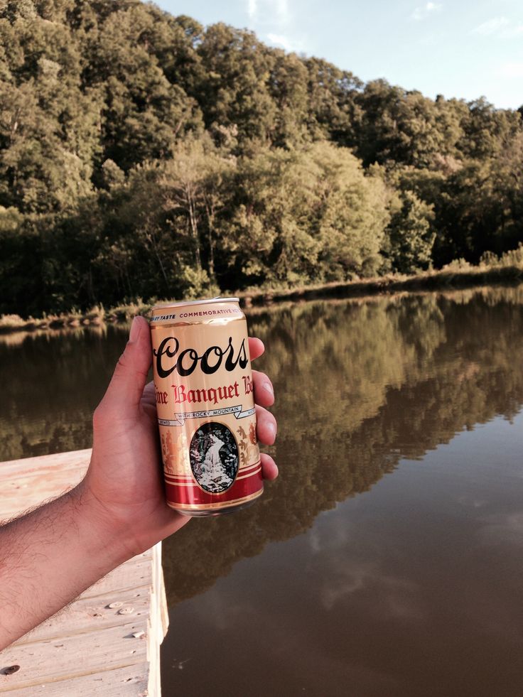 a hand holding a can of coors on a dock next to a body of water