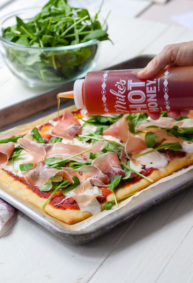 someone pouring ketchup onto a pizza with spinach and ham on the top