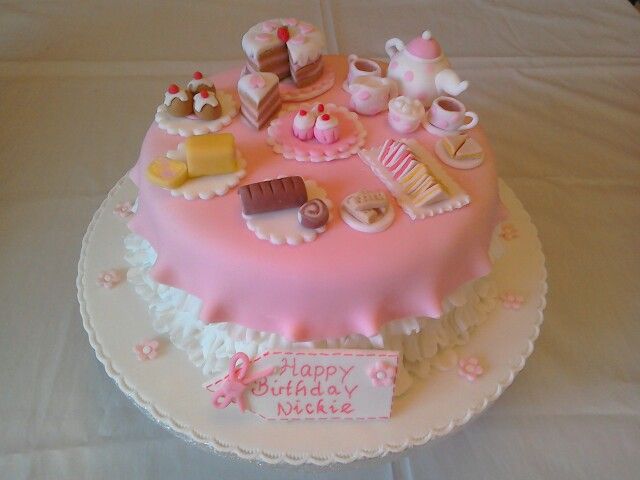 a pink birthday cake on a white tablecloth with tea and desserts around it