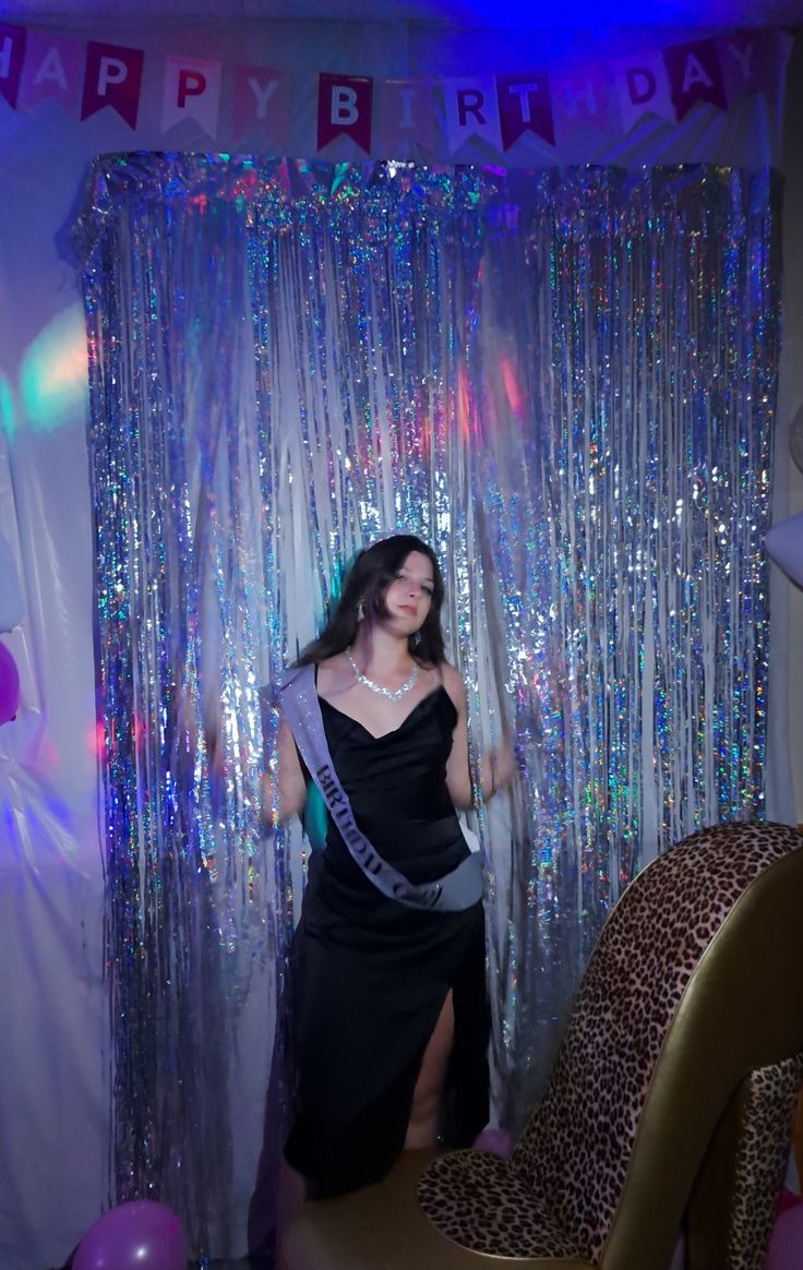 a woman in a black dress standing next to a chair and curtain with the words happy birthday on it