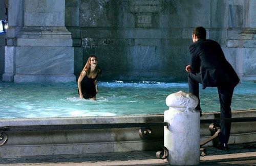 a man and woman standing in the water at a fountain, one is wearing a black suit