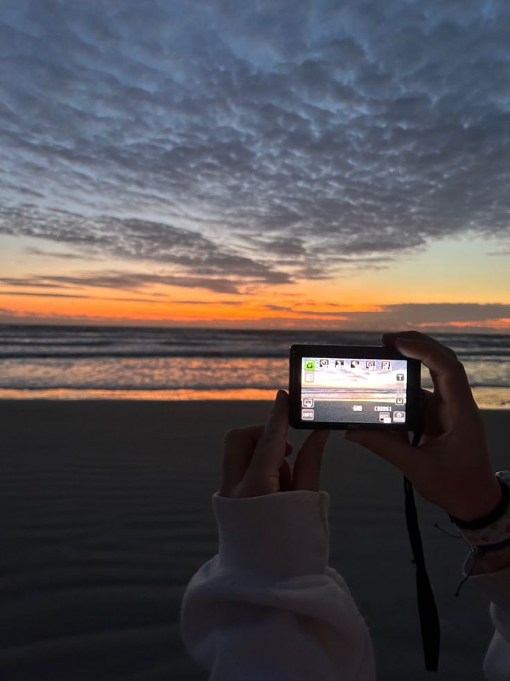 a person holding up a cell phone to take a photo on the beach at sunset