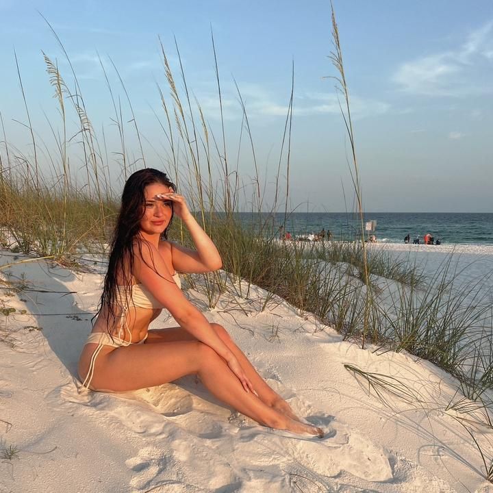 a beautiful woman sitting on top of a sandy beach next to tall grass and sea oats