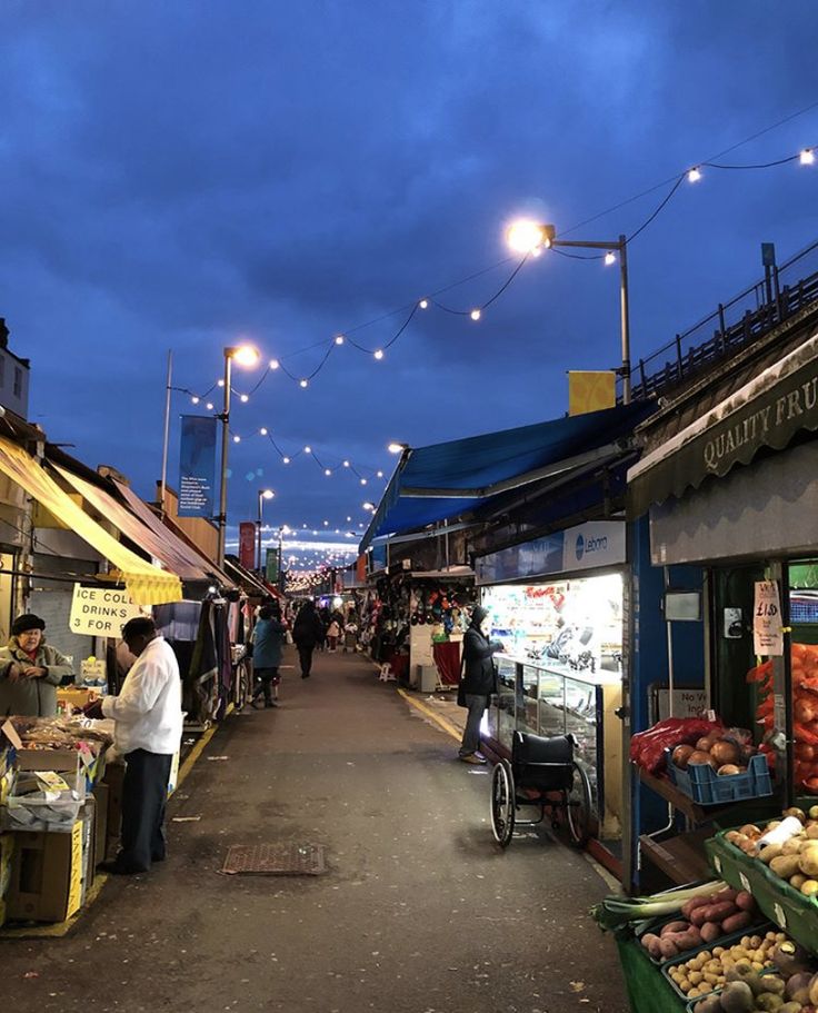 people are shopping at an outdoor market under the evening sky with street lights above them