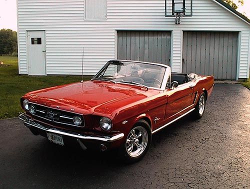 an old red mustang convertible parked in front of a white barn with two garage doors