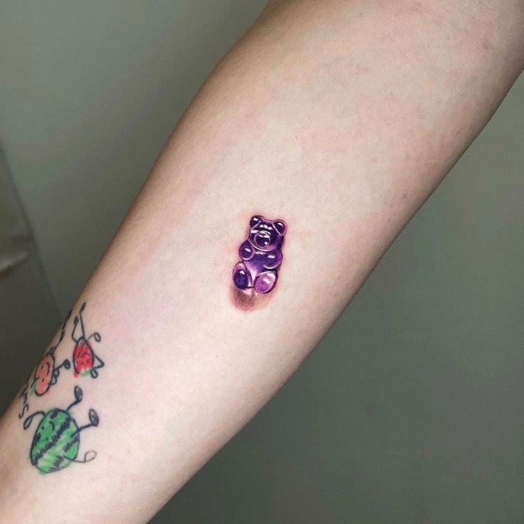 a small purple teddy bear tattoo on the right arm and wrist, next to an apple