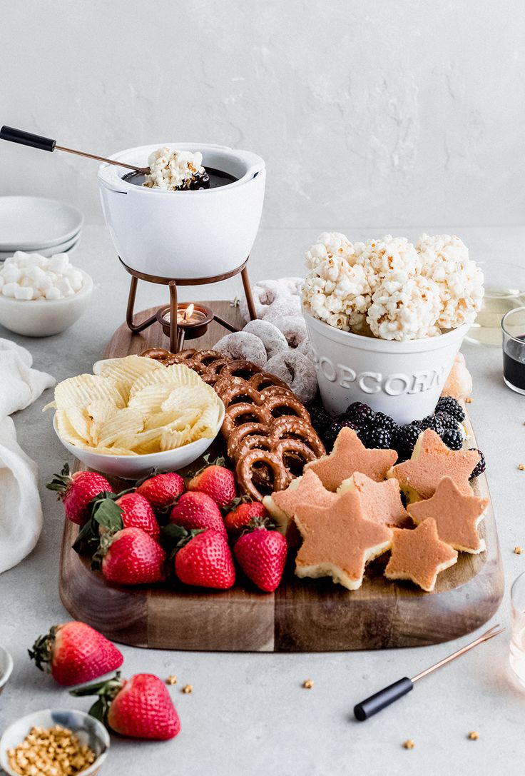 various snacks are arranged on a cutting board with bowls of popcorn, strawberries and cookies