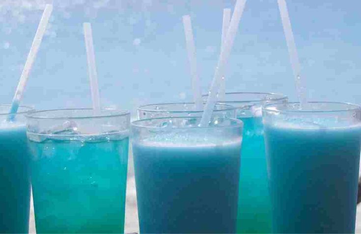 three glasses filled with blue liquid sitting on top of a table next to the ocean