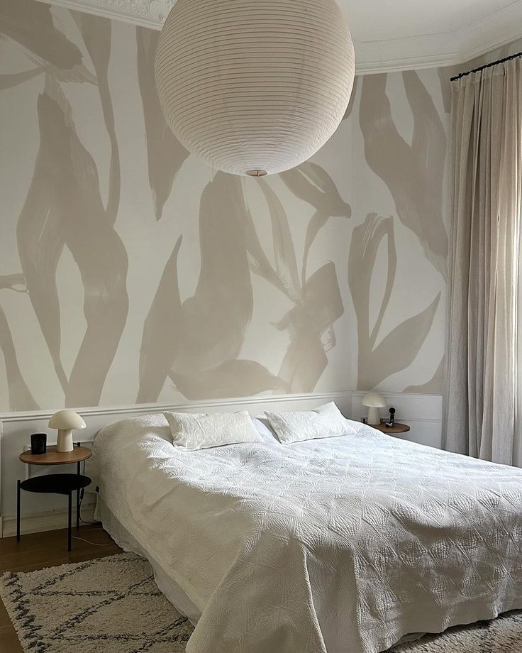 a white bed sitting under a light fixture in a bedroom next to a wall with leaves painted on it