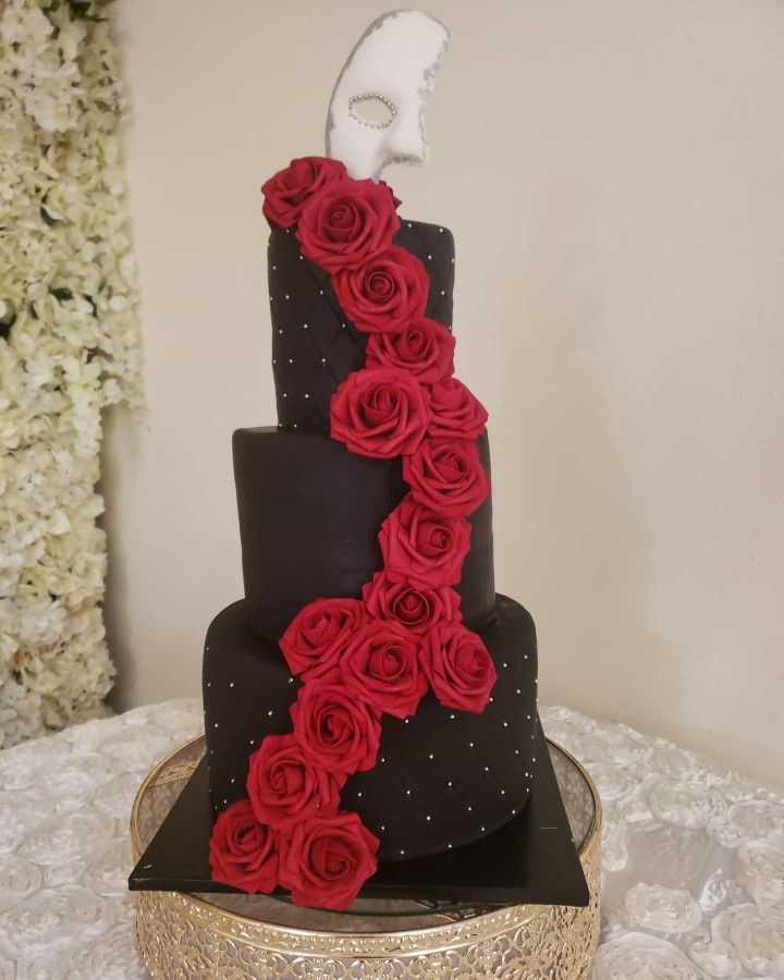 a three tiered black and red wedding cake with roses on the side, topped with a white mask