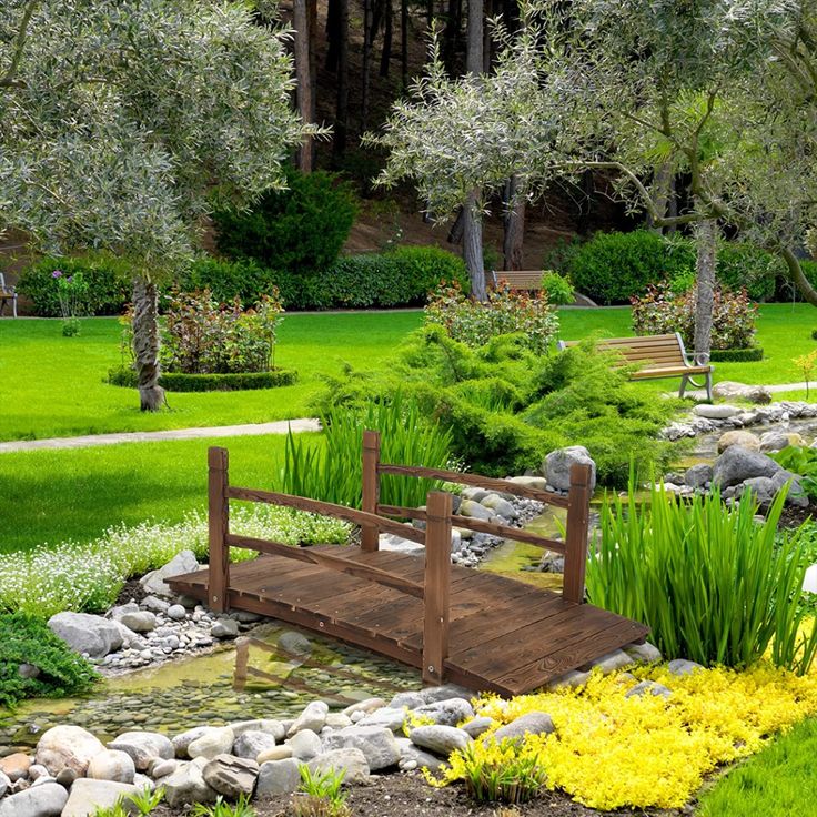 a wooden bridge over a small pond in a park with green grass and flowers around it