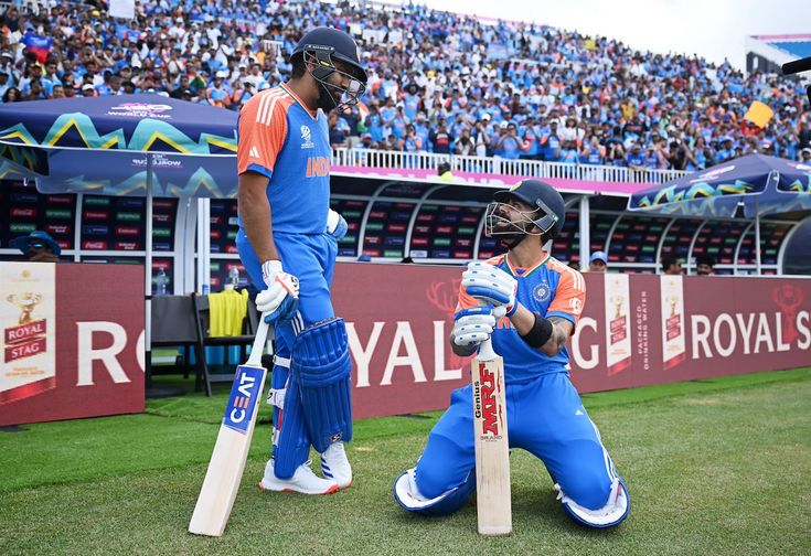 two men in blue and orange uniforms playing cricket on grass with spectators watching from the bleachers