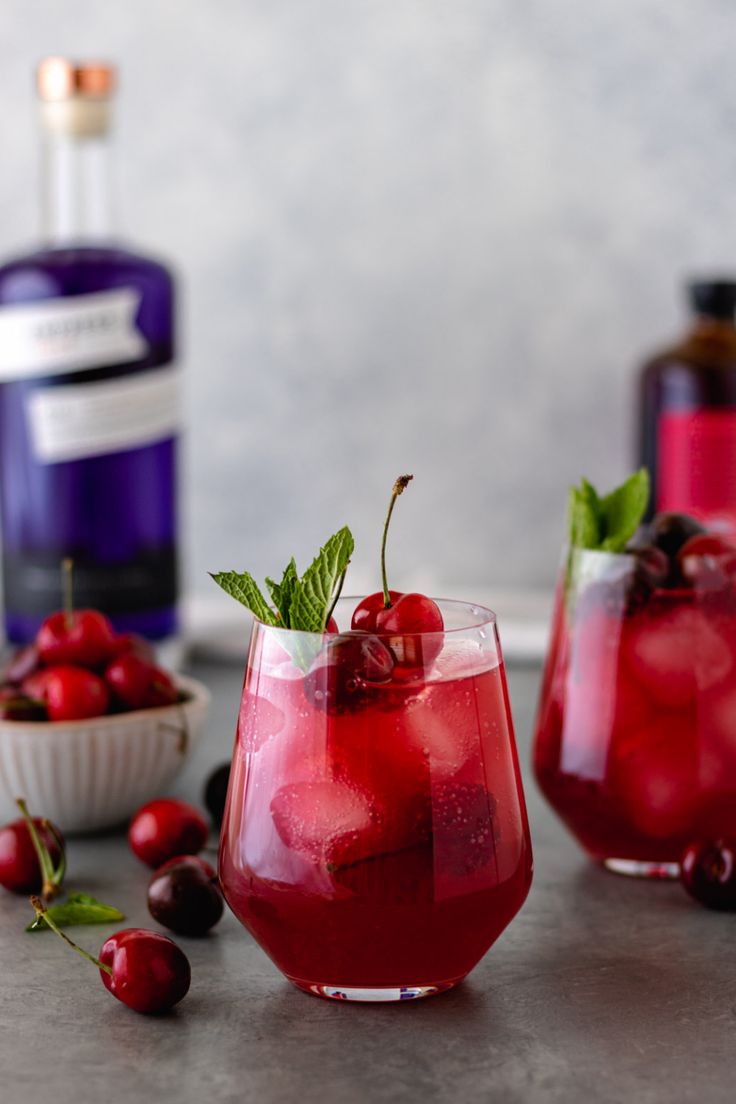 two glasses filled with red liquid and cherries next to a bowl of cherries