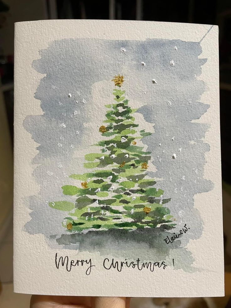 a hand holding up a christmas card with a watercolor painting of a pine tree