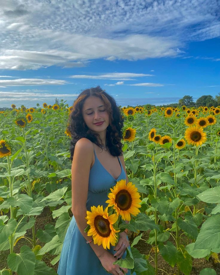 a woman in a blue dress holding sunflowers on a sunny day with clouds