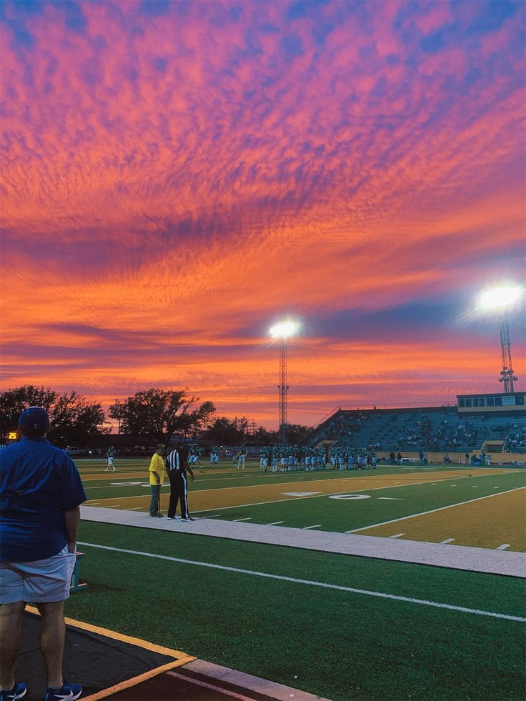 people are watching the sun set on a football field with lights in the distance and clouds in the sky