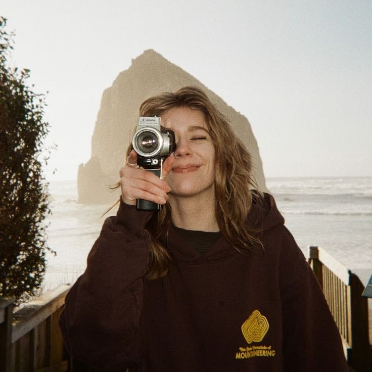 a woman taking a selfie with her camera near the ocean and mountains in the background