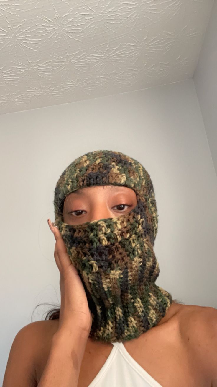 a woman wearing a camo knitted mask covering her face with one hand and looking at the camera