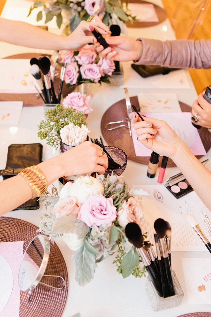 several people are sitting at a table with flowers and makeup brushes on it, all holding their hands together