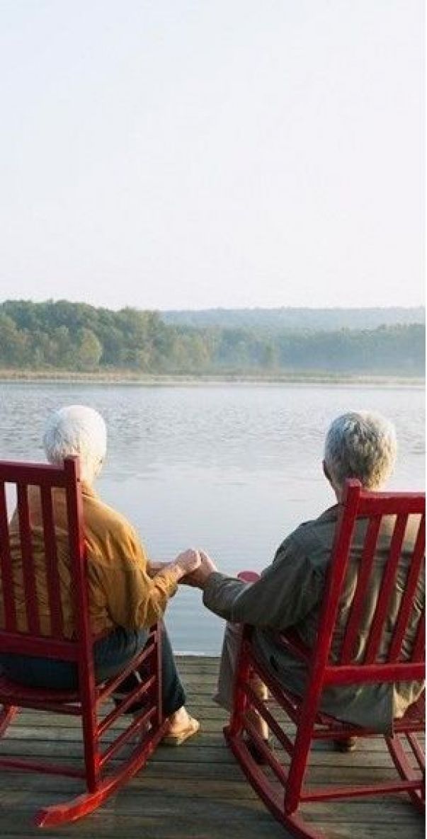 two elderly people sitting on red rocking chairs looking out at the water and trees in the distance