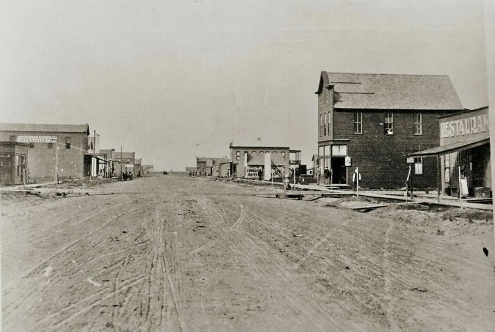 an old black and white photo of a dirt road in the middle of town with buildings on both sides