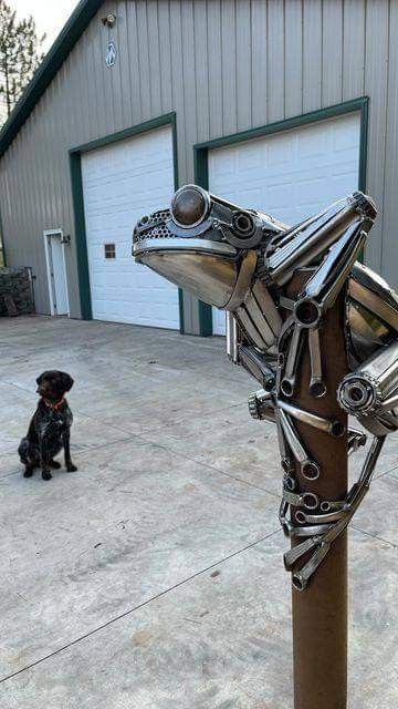 a dog sitting next to a metal frog sculpture on top of a wooden pole in front of a garage