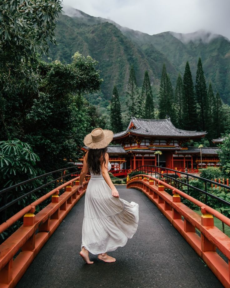 a woman in a white dress and straw hat walking across a bridge with mountains in the background