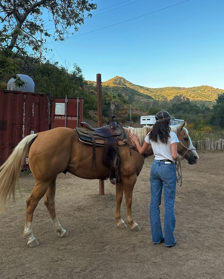 a woman standing next to a brown horse on top of a dirt field with mountains in the background