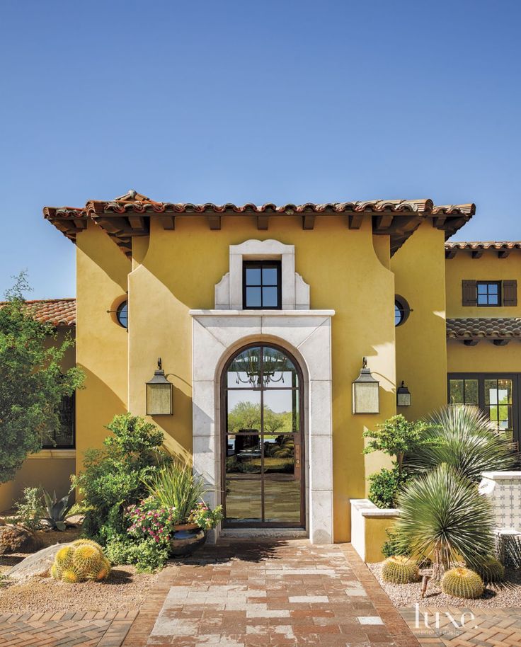 a yellow house with an arched doorway and brick walkway leading to the front door is surrounded by greenery