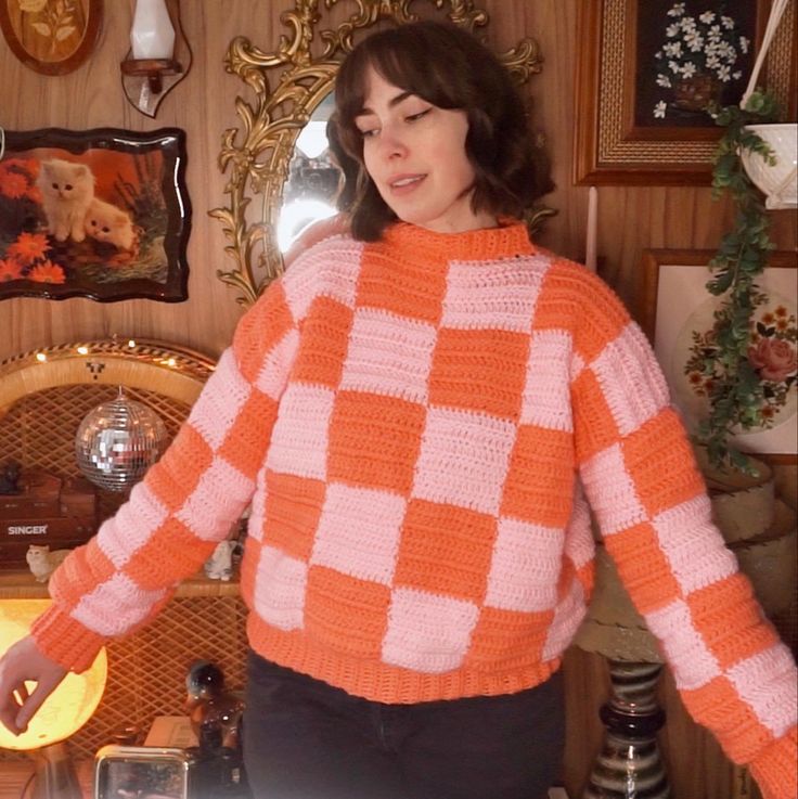 a woman wearing an orange and white sweater standing in front of a wall with pictures on it