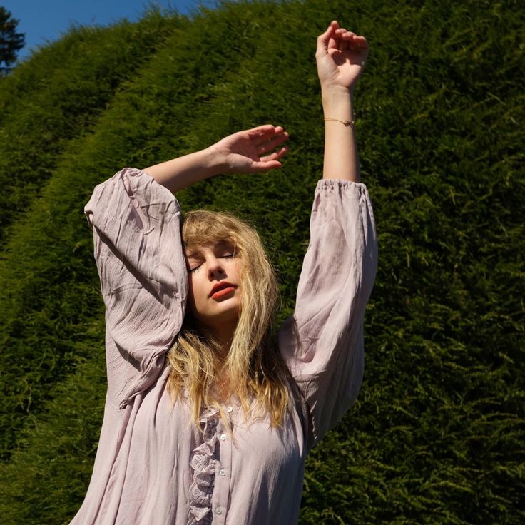 a woman standing in front of a lush green hillside holding her hands up to the sky