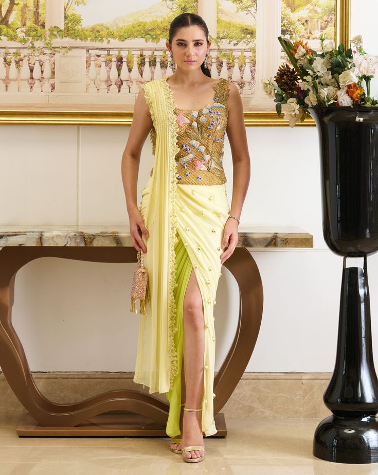 Rhyming perfectly with your day-time soirees, Lulu is a pre-stitched saree featuring a golden-hued designer blouse made out of fish-net fabric with floral embroidery detailing and a front pallu. The innovative draping style creates an exquisite piece immersed in the complimentary palette of lemon, presenting a fresh look for seasonal affairs. A perfect amalgamation of modernity and sophistication indulged in the traditional silhouette is a must-have festive essential that your wardrobe has been missing out on. Sleeveless blouse crafted out of fish-net fabric and adorned with beautiful floral embroidery detailing. The pre-draped skirt featuring a slit is adorned with dainty detailing of embroidered drops. Front pallu embellished with a linear sequined pattern and floral cut-work detailing a Sleeveless Pre-draped Saree With Resham Embroidery For Party, Embroidered Silk Pre-draped Saree For Evening, Evening Embroidered Pre-draped Saree, Festive Yellow Fitted Pre-draped Saree, Evening Embroidered Party Wear Pre-draped Saree, Evening Party Wear Embroidered Pre-draped Saree, Party Wear Embroidered Pre-draped Saree For Evening, Sleeveless Embroidered Pre-draped Saree For Party, Gold Pre-draped Saree With Resham Embroidery For Evening