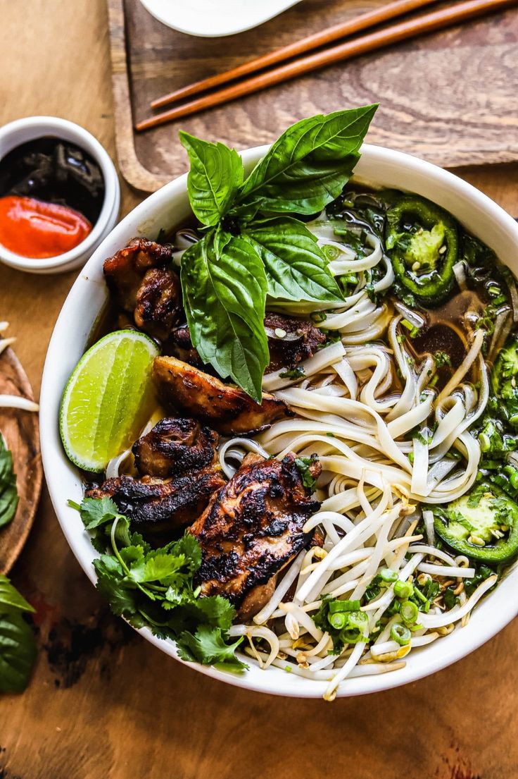 a white bowl filled with noodles, meat and veggies next to chopsticks