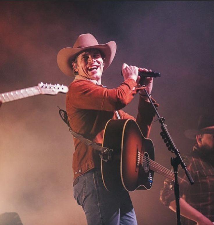 a man with a cowboy hat holding a guitar and singing into a microphone on stage