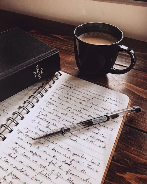 an open notebook and pen sitting on top of a wooden table next to a cup of coffee