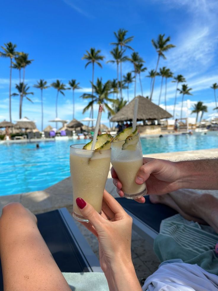 two people toasting with cocktails in front of an outdoor pool and palm trees